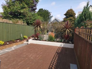 An outdoor garden area with a wooden deck bordered by green and brown fences and planted flowers at Hawthorn Cottage in St Teath