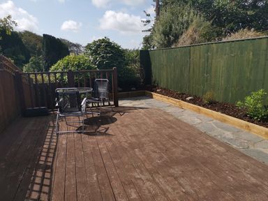 An outdoor wooden deck with a glass table and three metal chairs next to a green wooden fence and garden beds at Hawthorn Cottage in St Teath