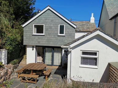 A backyard with a wooden picnic table and benches next to a two-story gray and white house at Hawthorn Cottage in St Teath