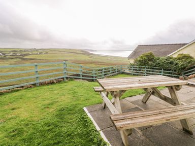 A wooden picnic table and benches on a patio with a grass yard and views of fields and coastline at Valley High in Solva