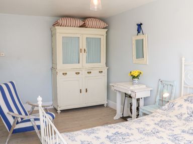 A bedroom with a white cabinet topped with pillows a blue and white striped chair a small table with flowers and a bed with floral bedding at 3 Elm Cottage in Carhampton