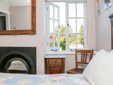 A bedroom with an open window a wooden chair a bedside cabinet and a bed with a patterned quilt at 3 Elm Cottage in Carhampton