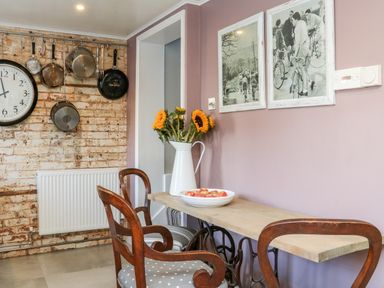 A kitchen corner with a wooden table and two chairs sunflowers in a white jug and a bowl of apples on the table pots and pans hanging on a brick wall with a large clock at 3 Elm Cottage in Carhampton