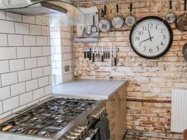 A kitchen with a gas stove a countertop hanging pans and knives on a brick wall and a large clock at 3 Elm Cottage in Carhampton