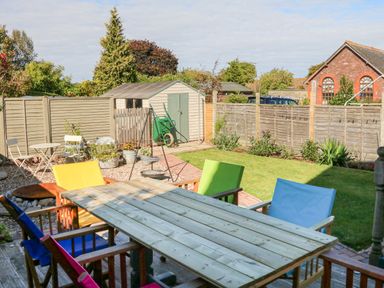 A backyard with a wooden table and colorful chairs on a deck a lawn shed and garden plants at 3 Elm Cottage in Carhampton