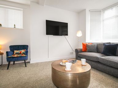 A living room with a wall-mounted TV a blue armchair and a grey sofa with colorful cushions at The White Cottage in Weymouth