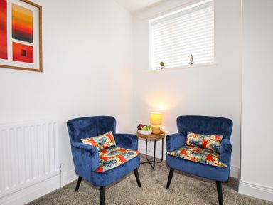 A seating area with two blue armchairs with floral cushions a small round wooden table with a lamp and fruit bowl a window with blinds and two small plants on the sill at The White Cottage in Weymouth