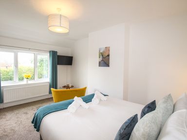 A bedroom with a double bed with pillows and rolled towels a yellow chair a small round table a wall-mounted TV and a window with blue curtains at The White Cottage in Weymouth