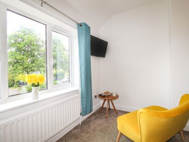 A room with a yellow chair a small round wooden table a wall-mounted television and a window with blue curtains at The White Cottage in Weymouth