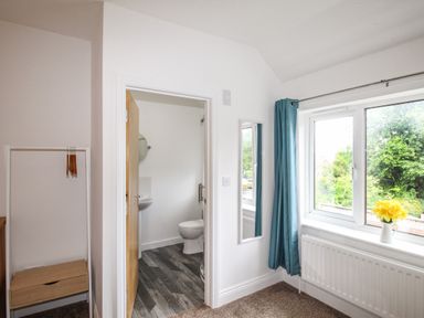 A room with a window blue curtains and a small wooden drawer unit next to a doorway leading to a bathroom at The White Cottage in Weymouth