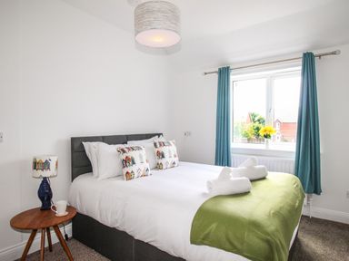 A bedroom with a double bed next to a window with blue curtains and a wooden side table with a lamp and a cup at The White Cottage in Weymouth