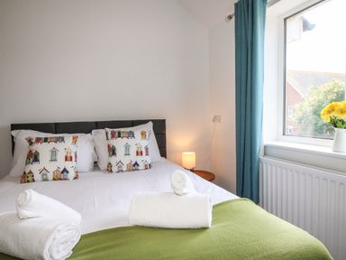 A bedroom with a double bed with colorful cushions white towels on a green blanket next to a window with blue curtains and a vase of yellow flowers at The White Cottage in Weymouth