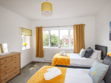 A bedroom with two single beds with yellow throws and pillows a wooden chest of drawers and windows with yellow curtains at The White Cottage in Weymouth