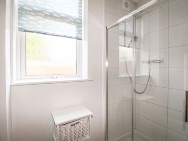 A bathroom with a window with a fish patterned blind a white wicker basket and a glass shower enclosure at The White Cottage in Weymouth