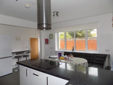 A kitchen with a black countertop island, stove, bar stools, refrigerator, table with items, corner seating and large windows at The White Cottage in Weymouth