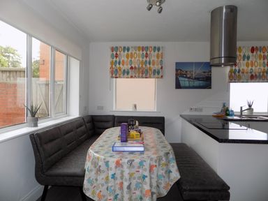 A kitchen dining area with a table covered by a patterned tablecloth surrounded by black cushioned benches at The White Cottage in Weymouth