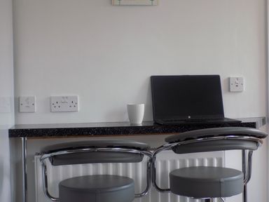 A workspace with a black laptop and a white cup on a black counter with two gray cushioned stools at The White Cottage in Weymouth