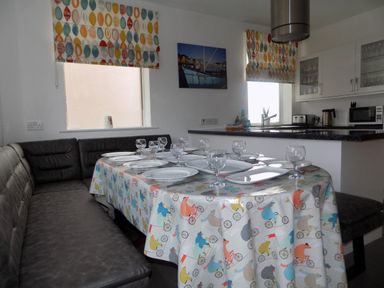 A dining area with a table set with plates and glasses next to a gray cushioned bench and a kitchen with appliances and windows with colorful blinds at The White Cottage in Weymouth