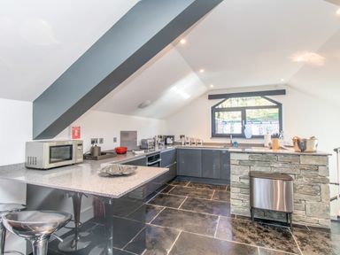 A kitchen with gray cabinets a microwave a stone island and bar stools at The Boat House at Louper Weir Bowness-On-Windermere