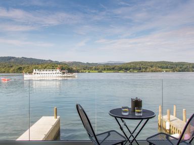 A small black table and two chairs on a balcony overlooking a lake with a boat and distant hills at The Boat House at Louper Weir in Bowness-On-Windermere