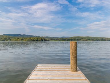 A wooden dock extending into a calm lake with forested hills in the background at The Boat House at Louper Weir in Bowness-On-Windermere
