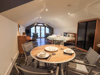 A room with a round dining table set for two, a bed, two chairs facing a window, and a wardrobe at The Boat House at Louper Weir in Bowness-On-Windermere
