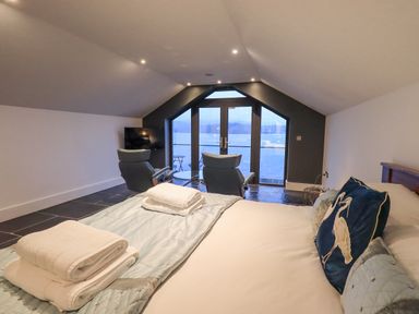 A bedroom with a bed pillows and towels two chairs a television and glass doors overlooking water at The Boat House at Louper Weir Bowness-On-Windermere