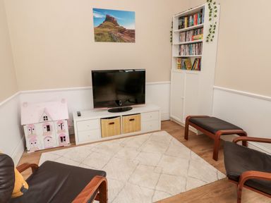 A living room with a television on a stand a white cabinet with books two chairs a dollhouse and a rug on the floor at The Old Sweet Shop in Alnwick