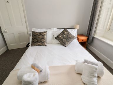 A bedroom with a bed with white linens two patterned pillows a wooden nightstand with a lamp towels on the bed and a window with curtains at The Old Sweet Shop in Alnwick