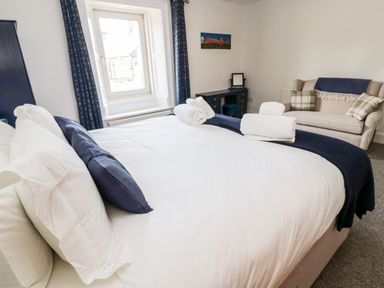 A bedroom with a bed with white and blue pillows a window with curtains a desk and a beige sofa with cushions at The Old Sweet Shop in Alnwick