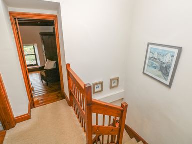 A staircase with wooden railing and framed pictures on the walls next to a doorway leading to a room with an armchair at Inglenook Cottage in Broad Haven