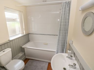 A bathroom with a bathtub shower combination a toilet a sink with two taps and a round mirror at Inglenook Cottage in Broad Haven