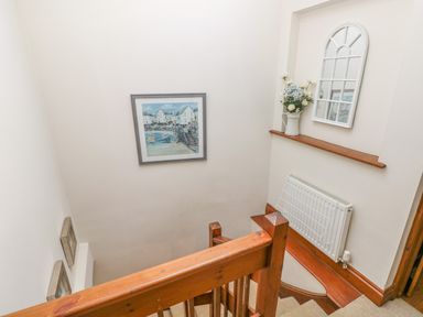 A stairway with wooden railing a framed painting on the wall a white radiator a shelf with a vase of flowers and a decorative window mirror at Inglenook Cottage in Broad Haven