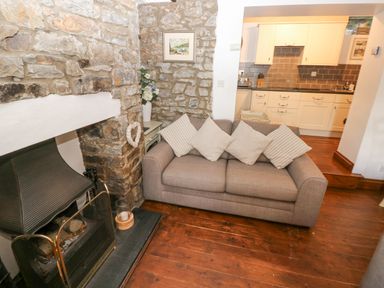 A living room with a gray sofa and cushions next to a stone fireplace with a fireguard and an open view of a kitchen at Inglenook Cottage in Broad Haven