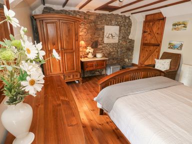 A bedroom with wooden furniture including a bed wardrobe chair and dresser with flowers on the dresser at Inglenook Cottage in Broad Haven
