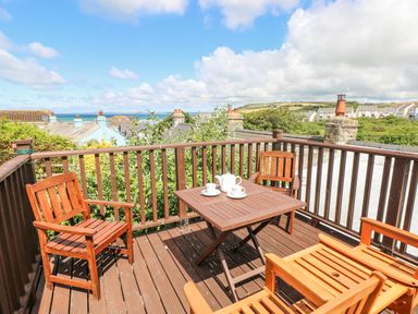 A wooden balcony with four wooden chairs and a wooden table with a teapot and cups overlooking rooftops and a distant coastline at Inglenook Cottage in Broad Haven