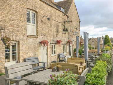 Outdoor seating area with wooden benches and tables beside a stone building with hanging plants and covered umbrellas at 3 Painters Field Quenington Near Cirencester