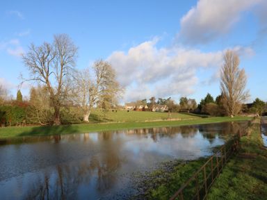 A pond with trees and houses in the background at 3 Painters Field Quenington Near Cirencester