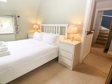 A bedroom with a white bed two bedside tables with lamps and a doorway showing stairs at 3 Painters Field Quenington Near Cirencester