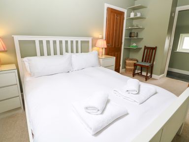 A bedroom with a white bed and white linens with towels on top beside wooden door and chair at 3 Painters Field in Quenington Near Cirencester