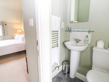 A small bathroom with a sink a toilet a towel rack a mirror and a bedroom with a bed and lamp at 3 Painters Field in Quenington Near Cirencester