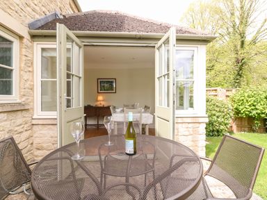 A round metal patio table with a bottle and two glasses outside open double doors leading to a dining room at 3 Painters Field Quenington Near Cirencester