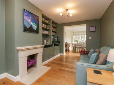 A living room with a fireplace a built in bookshelf a blue sofa and a dining room visible through a doorway at 3 Painters Field Quenington Near Cirencester