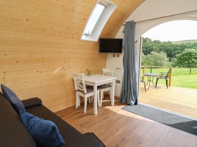 An interior room with wooden walls a small white dining table with two chairs a wall-mounted TV a black sofa with blue cushions and an open door leading to a deck with outdoor furniture and green landscape visible at Seren in Llandrindod Wells