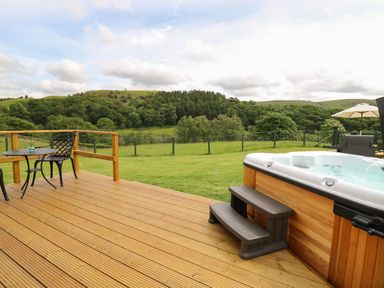 A wooden deck with a hot tub a small round table with two chairs and green hills in the background at Seren in Llandrindod Wells