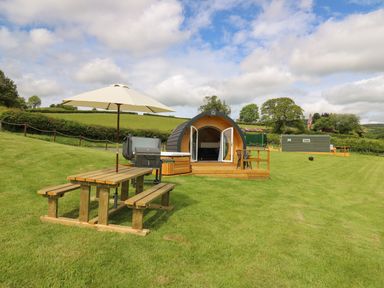 An outdoor campsite with a wooden picnic table with an umbrella barbecue grills two small wooden pods and green fields at Seren in Llandrindod Wells
