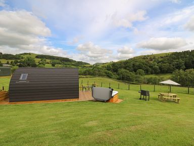 A small curved cabin with a deck hot tub a barbecue and a picnic table with umbrella in a grassy area with hills in the background at Seren in Llandrindod Wells