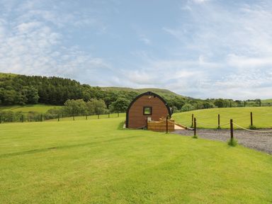 A small wooden cabin on a grassy field with hills and trees in the background at Seren in Llandrindod Wells