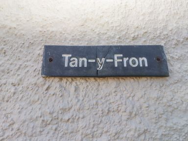 A cracked black nameplate with white letters mounted on a textured white wall at Tanyfron Holiday Studio in Bryngwran near Gwalchmai