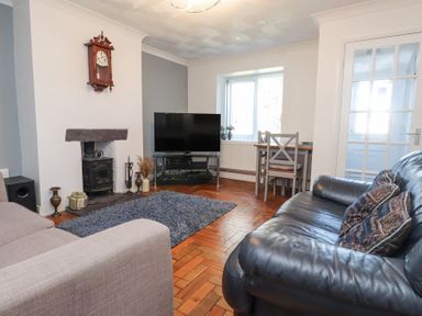 A living room with a black leather sofa a beige armchair a flat screen tv on a stand a small dining table with chairs and a wood burning stove at Tanyfron Holiday Studio in Bryngwran near Gwalchmai